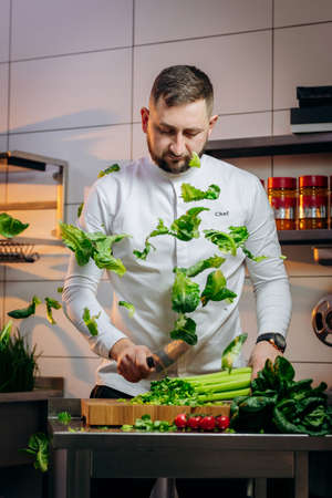 Portrait of handsome chef cooking salad in the kitchen. The cook cuts fresh green leaves of cabbage, spinach, arugula for preparing a vegetarian Salad. Concept of Cooking process and preparing dishes.の写真素材