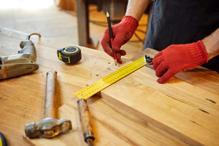 Carpenter working with a wood, marking plank with a pencil and taking measurements to cut a piece of...