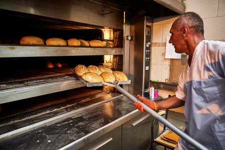 Professional baker in uniform takes out a cart with freshly baked bread from an industrial oven in a...
