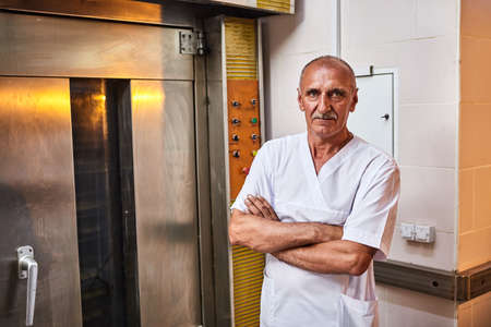 Portrait of professional baker in uniform stands on the background of industrial oven in a bakery...