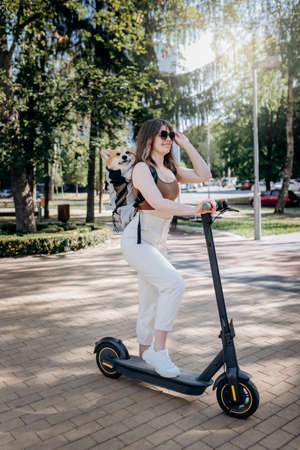 Happy smiling woman traveler is riding her electro scooter in city parkland with dog Welsh Corgi Pembroke in a special backpackの写真素材