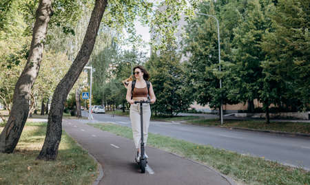 Happy smiling woman traveler is riding her electro scooter in city parkland with dog Welsh Corgi Pembroke in a special backpackの写真素材