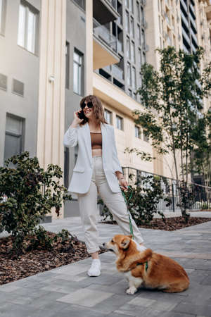 Attractive Woman wearing white suit and sunglasses walking down the street with Welsh Corgi Pembroke dog and talking phoneの写真素材