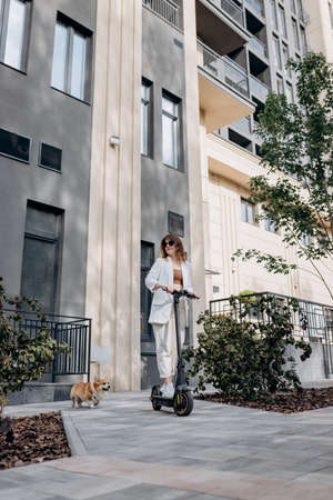 Beautiful young woman in sunglasses and white suit is riding on her electric scooter with Corgi dog near modern building in city and looking awayの写真素材