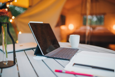 Digital tablet and notebook lying on table on background of Cozy open glamping tent with light inside during dusk. Luxury camping tent for outdoor summer holiday and vacation. Lifestyle conceptの写真素材