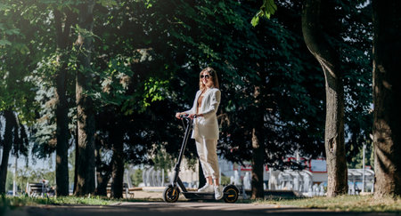 Beautiful young woman in sunglasses and white suit is standing with her electric scooter in city...