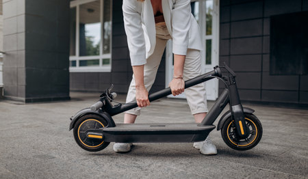 Close up of woman in white suit folding her electro scooter after ride while standing on background of modern buildingの写真素材