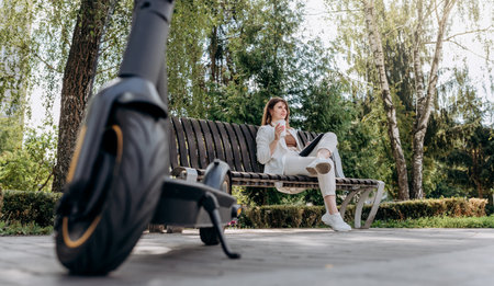 Pretty business woman in a white suit sits in a city park, drinks coffee and works on a digital tablet with an electric scooter in the foregroundの写真素材