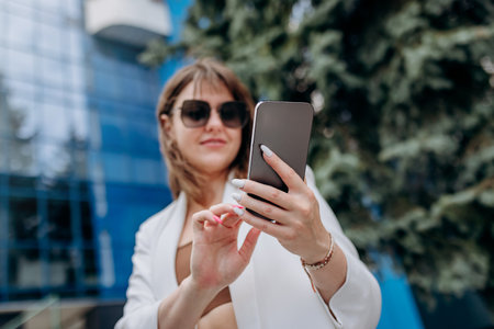 Smiling business woman in white suit and sunglasses using phone during break standing near modern office buildingの写真素材