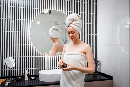Young attractive woman taking moisturizing cream from bottle in bathroom. Girl taking care of her dry complexionの写真素材