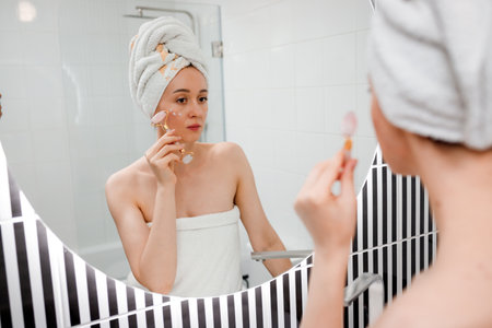 Beautiful woman with perfect skin wearing white towel after shower, making face massage using a jade face roller with natural quartz stone in bathroom. Natural cosmetics concept, wrinkle smoothingの写真素材