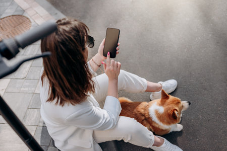 Top view of businesswoman in white suit sitting on electric scooter and using phone during walking with Welsh Corgi Pembroke dog in cityの写真素材