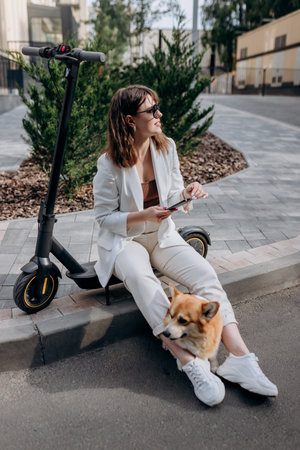 Young businesswoman in white suit sitting on electric scooter and using phone during walking with Welsh Corgi Pembroke dog in cityの写真素材