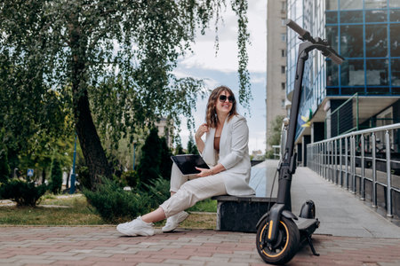 Smiling business woman in white suit and sunglasses working on digital tablet sitting near modern office building with electric scooter in the foregroundの写真素材