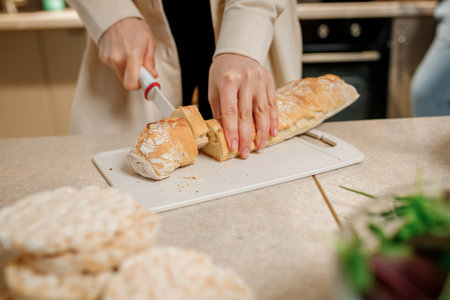 Close up of hands of a young woman cooks in her home kitchen, cuts a baguette bread with a knife