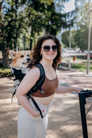 Happy smiling woman traveler is riding her electro scooter in city parkland with dog Welsh Corgi...