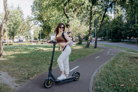 Happy smiling woman traveler is riding her electro scooter in city parkland with dog Welsh Corgi Pembroke in a special backpackの写真素材