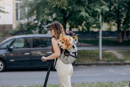 Happy smiling woman traveler is riding her electro scooter in city parkland with dog Welsh Corgi Pembroke in a special backpackの写真素材