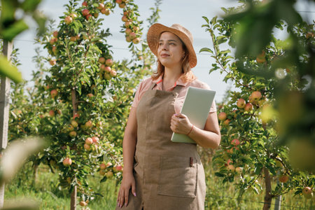 Attractive female agronomist or farmer with laptop standing in apple orchard and checking fruit, makes notes. Agriculture and gardening concept. Healthy nutritionの写真素材