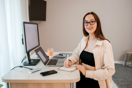Attractive business woman wearing casual clothes working at home office on laptop and making notes. Young happy female student studying in online universityの写真素材