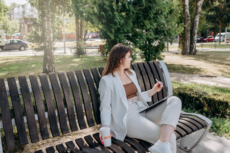 Pretty business woman in a white suit sits in a city park, drinks coffee and works on a digital...