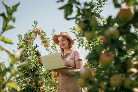 Attractive female agronomist or farmer with laptop standing in apple orchard and checking fruit, makes notes. Agriculture and gardening concept. Healthy nutritionの写真素材