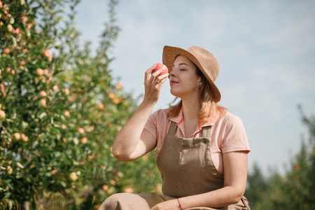 Happy smiling female farmer worker crop picking and smelling fresh ripe apples in orchard garden...