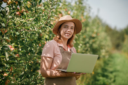 Attractive female agronomist or farmer with laptop standing in apple orchard and checking fruit, makes notes. Agriculture and gardening concept. Healthy nutritionの写真素材