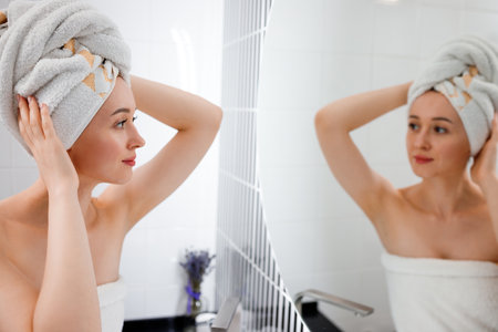 Attractive young woman in towel is looking at mirror standing in bathroom after taking shower....