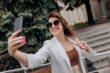 Smiling business woman in white suit and sunglasses making video call to friend using phone during break standing near modern office buildingの写真素材