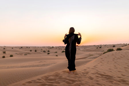 Back view of Beautiful mysterious woman in traditional arabic black long dress stands in the desert on sunsetの写真素材