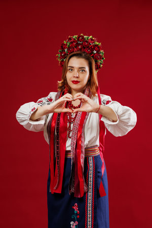 Ukrainian woman in traditional ethnic clothing and floral red wreath on viva magenta studio background with hands showing a heart shape. Ukrainian national embroidered dress call vyshyvankaの写真素材