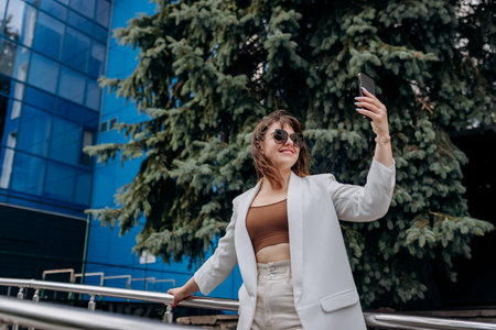 Smiling business woman in white suit and sunglasses making video call to friend using phone during break standing near modern office buildingの写真素材