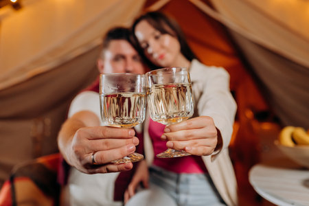 Happy lovely couple relaxing in glamping on summer evening and drinking wine near cozy bonfire. Luxury camping tent for outdoor recreation and recreation. Lifestyle conceptの写真素材