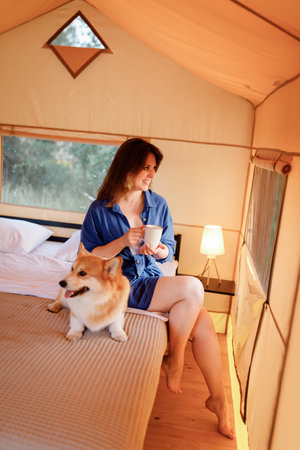Happy young woman with Welsh Corgi Pembroke dog relaxing and drink coffee in glamping on summer day. Luxury camping tent for outdoor recreation and recreation. Lifestyle conceptの写真素材