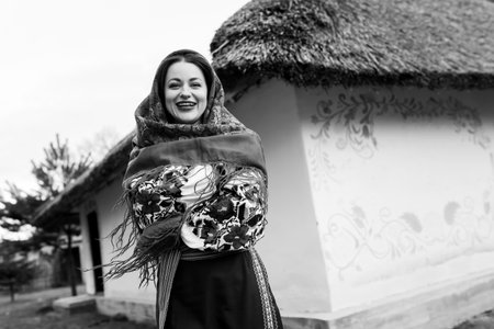 Charming woman in traditional ukrainian handkerchief, necklace and embroidered dress standing at background of decorated hut. Ukraine, style, folk, ethnic cultureの写真素材