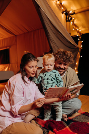 Happy family with lovely baby playing and read book together in glamping on summer evening near cozy bonfire. Luxury camping tent for outdoor recreation and recreation. Lifestyle conceptの写真素材