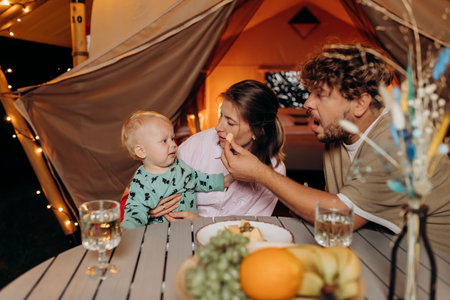 Happy family with lovely baby have dinner and spend time together in glamping on summer evening near cozy bonfire. Luxury camping tent for outdoor recreation and recreation. Lifestyle conceptの写真素材