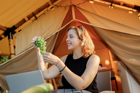 Smiling Woman freelancer eating grape while working on laptop in cozy glamping tent in a sunny day. Luxury camping tent for outdoor summer holiday and vacation. Lifestyle conceptの写真素材