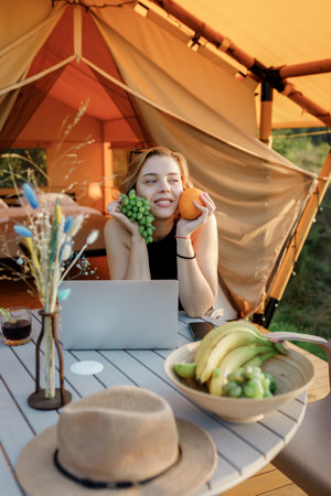 Smiling Woman freelancer holding grape and orange while working on laptop in cozy glamping tent in a...