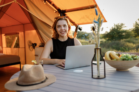 Smiling Woman freelancer using a laptop on a cozy glamping tent in a sunny day. Luxury camping tent for outdoor summer holiday and vacation. Lifestyle conceptの写真素材