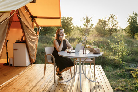 Smiling Woman freelancer talking by phone while working in cozy glamping tent in a sunny day. Luxury camping tent for outdoor summer holiday and vacation. Lifestyle conceptの写真素材