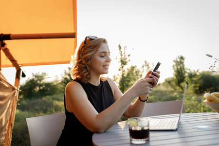 Smiling Woman freelancer using phone while working on laptop in cozy glamping tent in a sunny day. Luxury camping tent for outdoor summer holiday and vacation. Lifestyle conceptの写真素材