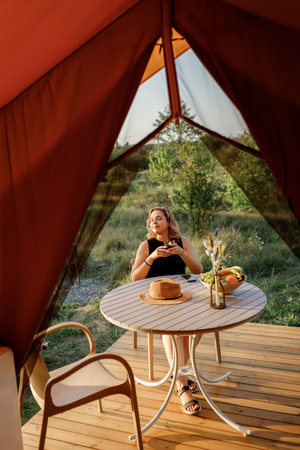 Happy Woman freelancer use phone and drinking cocktail call while relaxing on cozy glamping tent in sunny day. Luxury camping tent for outdoor summer holiday and vacation. Lifestyle conceptの写真素材