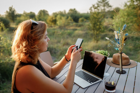 Smiling Woman freelancer using phone while working on laptop in cozy glamping tent in a sunny day. Luxury camping tent for outdoor summer holiday and vacation. Lifestyle conceptの写真素材