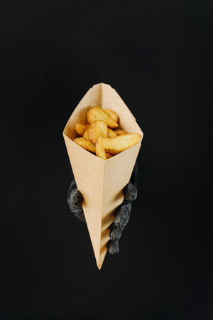 Hand holding French fries through torn holes in black paper background. Street food concept