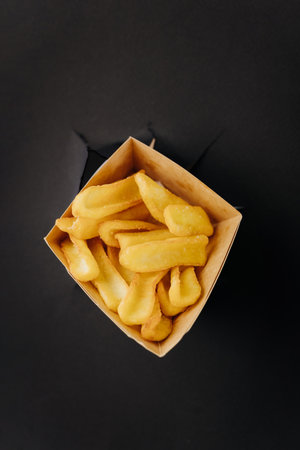 French fries sticks out through torn holes in black paper background. Street food concept