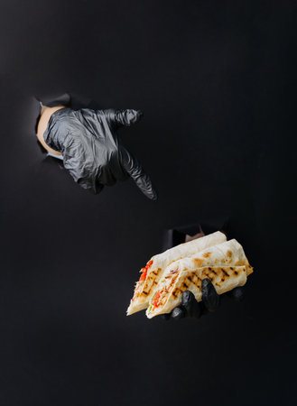 Hand holding doner or shawarma and point finger on it through torn holes in black paper background