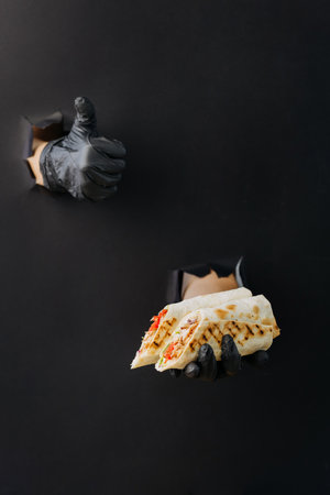 Hand holding doner and showing thumb up through torn holes in black paper backgroundの写真素材