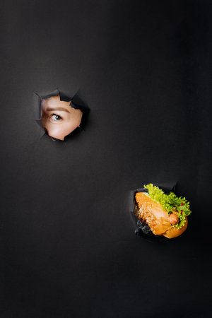 Female hand holding hot dog and looks through holes in black paper background. Street food conceptの写真素材